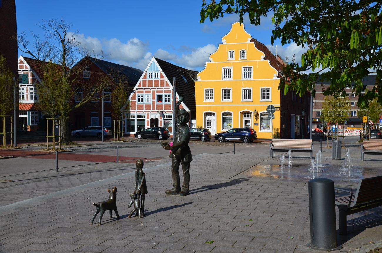 Kirchplatz square in Otterndorf with a red-and-white half-timbered house, a yellow gabled house, a bronze figure group in the foreground and water fountains behind