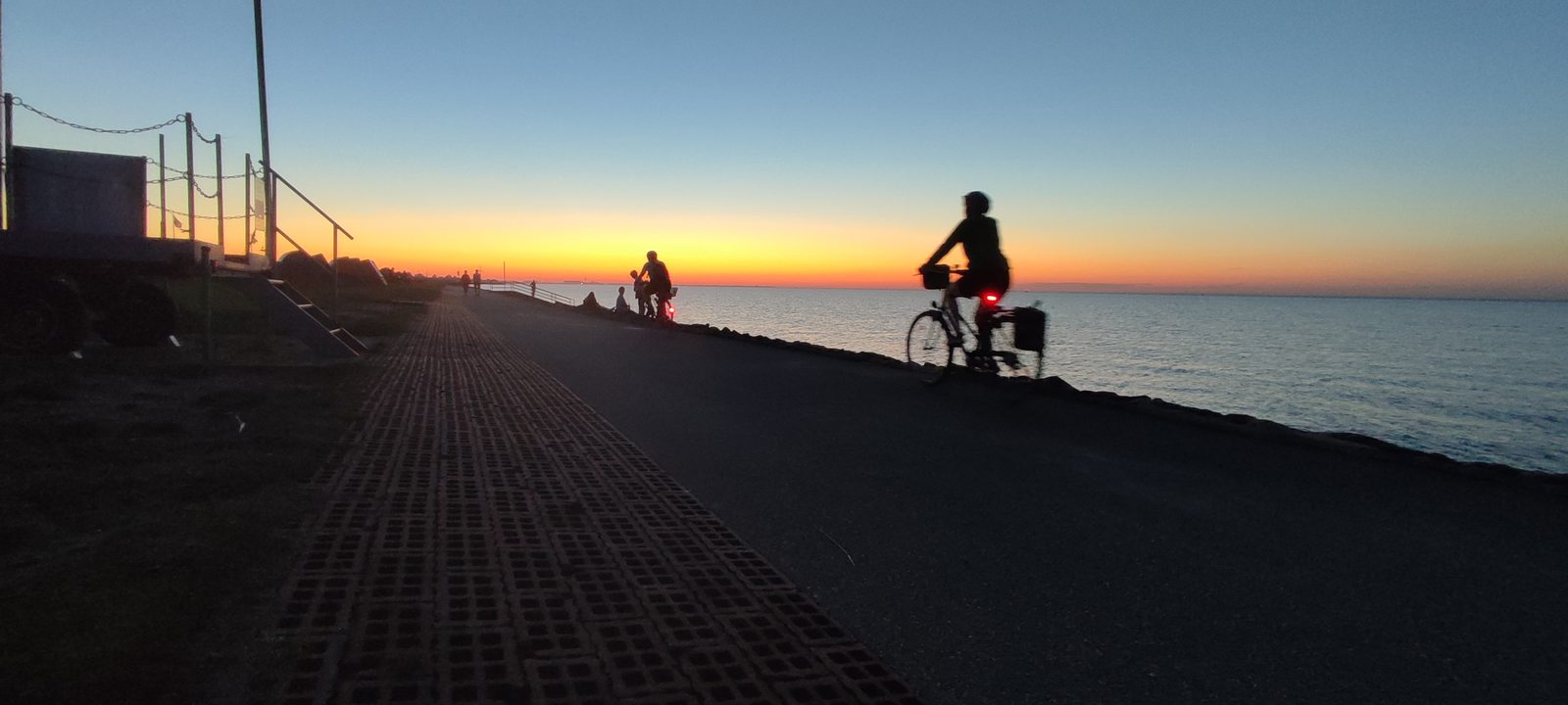 Radfahrer als Silhouette auf dem asphaltierten Deichweg an der Elbe in Otterndorf, orange leuchtender Horizont nach Sonnenuntergang, rechts das Wasser