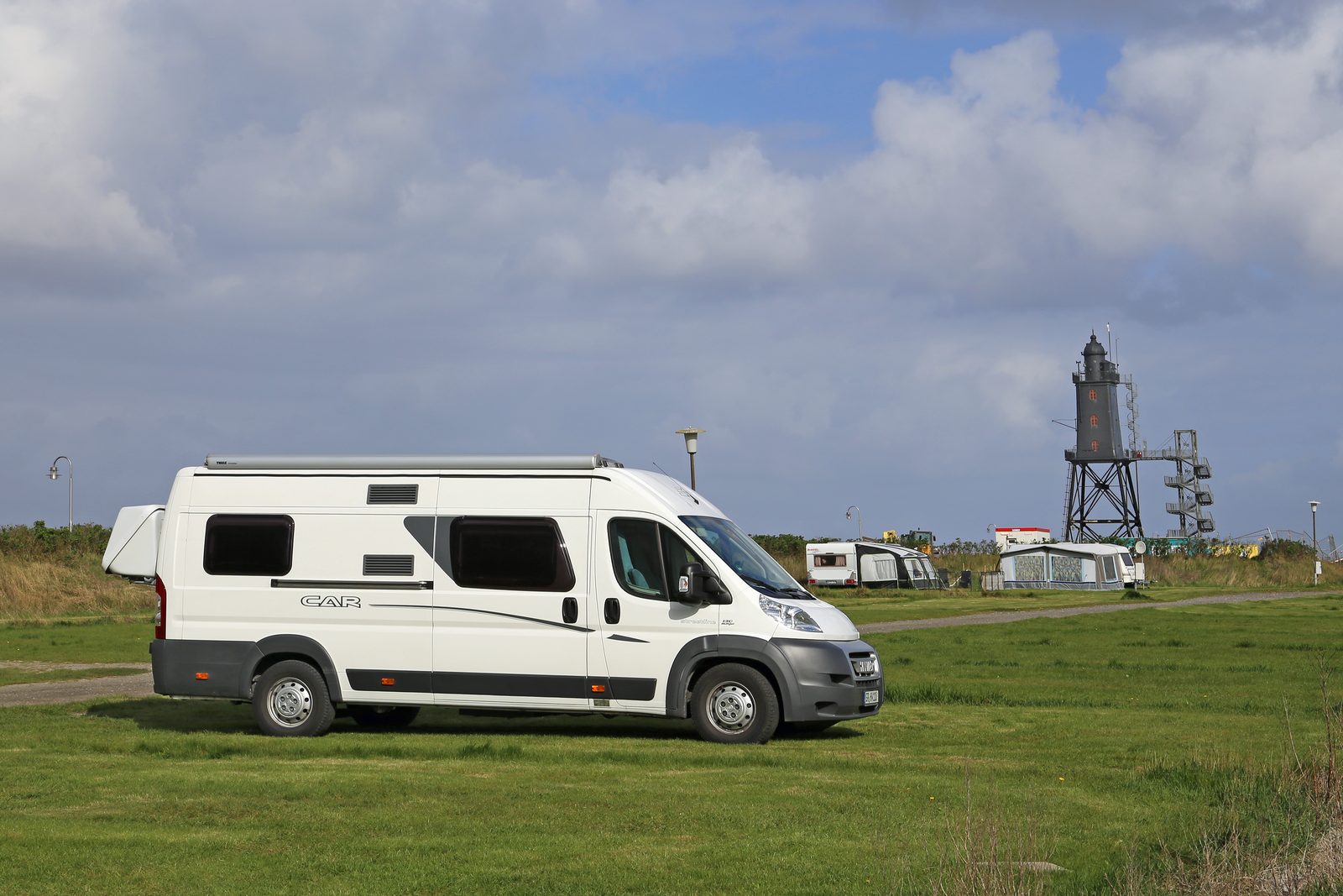 A white camper van on a grass pitch, with caravans and awnings behind it and the steel-framed Obereversand lighthouse in the distance