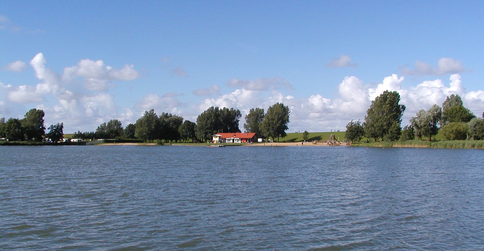 The See Achtern Diek lake in Otterndorf: a wide stretch of water with a sandy shore, trees and a red-roofed building behind it