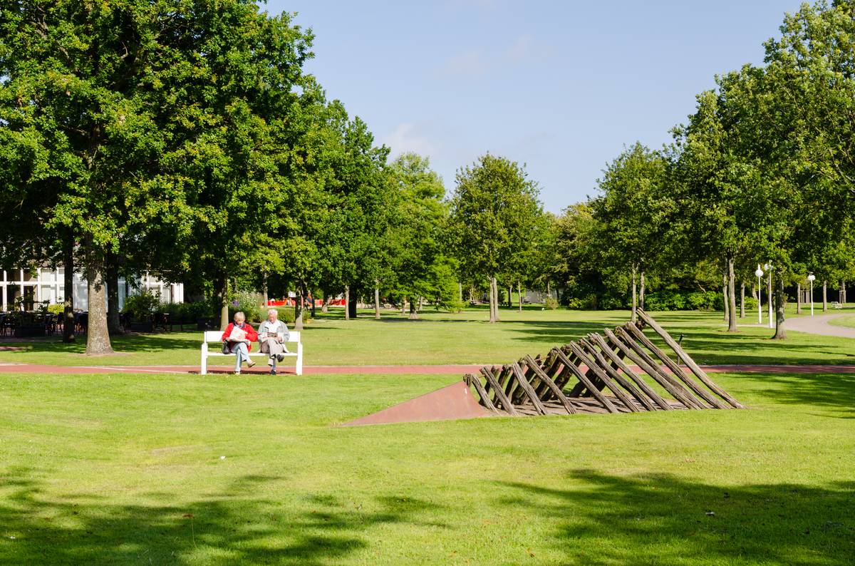 Weitläufige Rasenfläche im Kurpark Cuxhaven mit altem Baumbestand, einer weißen Parkbank mit zwei sitzenden Personen und einer liegenden Holzskulptur auf der Rasenfläche rechts