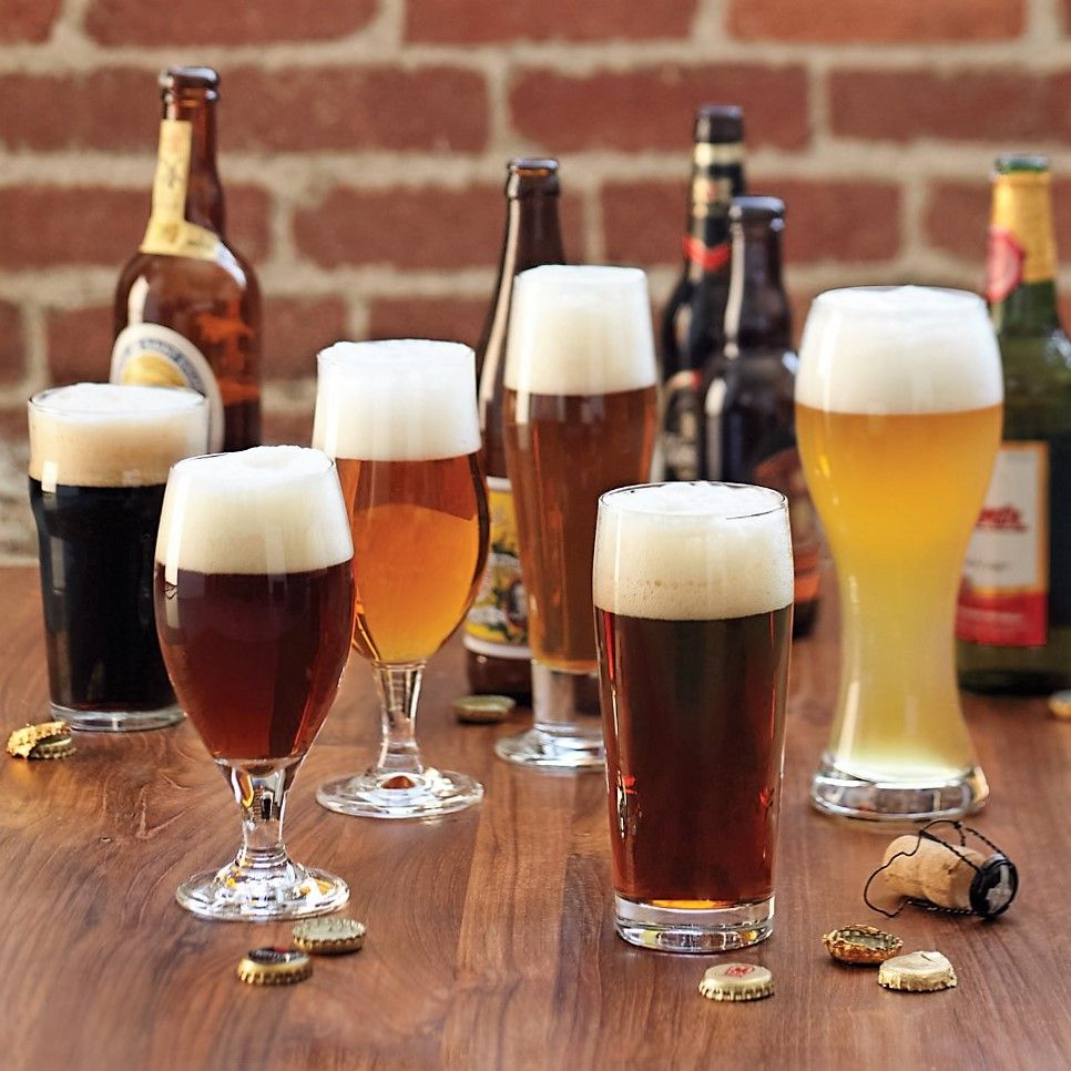 Sechs gefüllte Biergläser unterschiedlicher Form mit hellem und dunklem Bier, dahinter mehrere Bierflaschen vor einer Backsteinwand, davor Kronkorken und ein Korken auf einem Holztisch