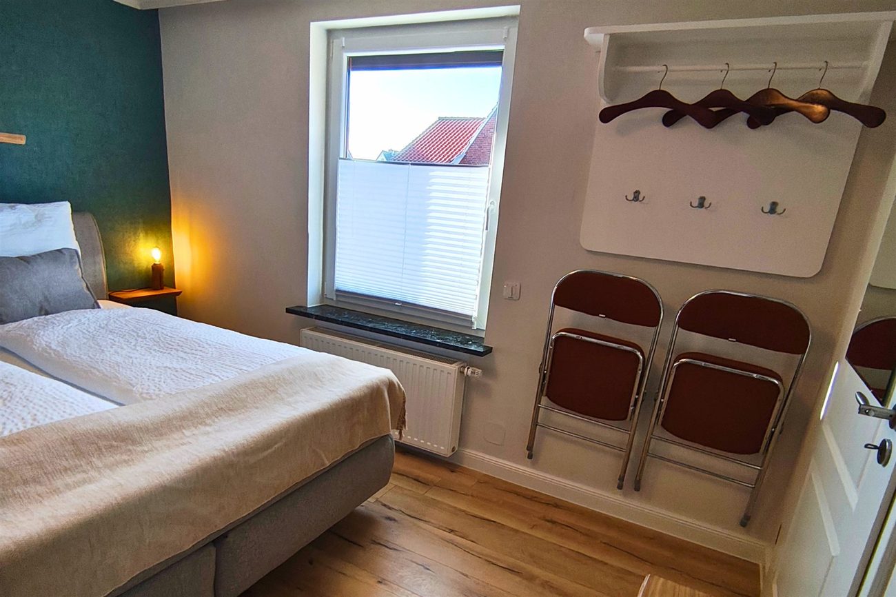Double bed against a petrol-blue feature wall in the Koje room, with a window looking out over red-tiled rooftops and a clothes rail with hangers beside it