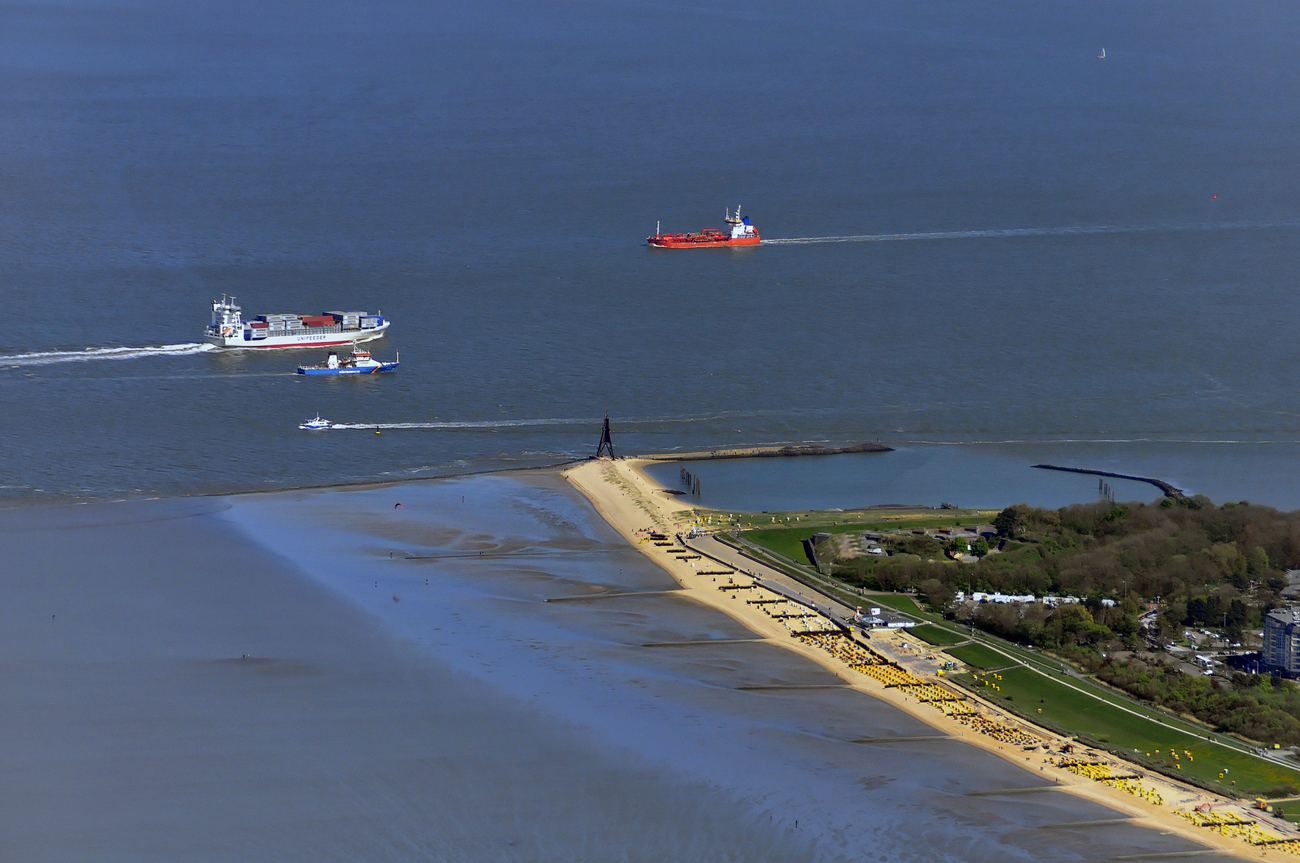 Luftaufnahme der Kugelbake bei Cuxhaven an der Spitze einer schmalen Sandnehrung, davor mehrere Schiffe auf der Elbe, dahinter Strand und eine breite grüne Fläche