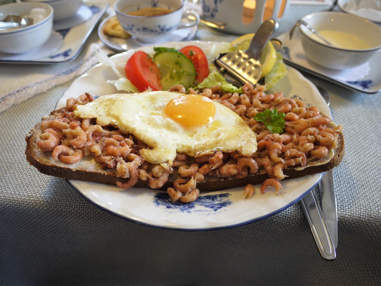 A slice of dark bread thickly topped with peeled shrimp, a fried egg on top, next to it slices of tomato, cucumber, lettuce and a slice of lemon on a white plate with a blue pattern