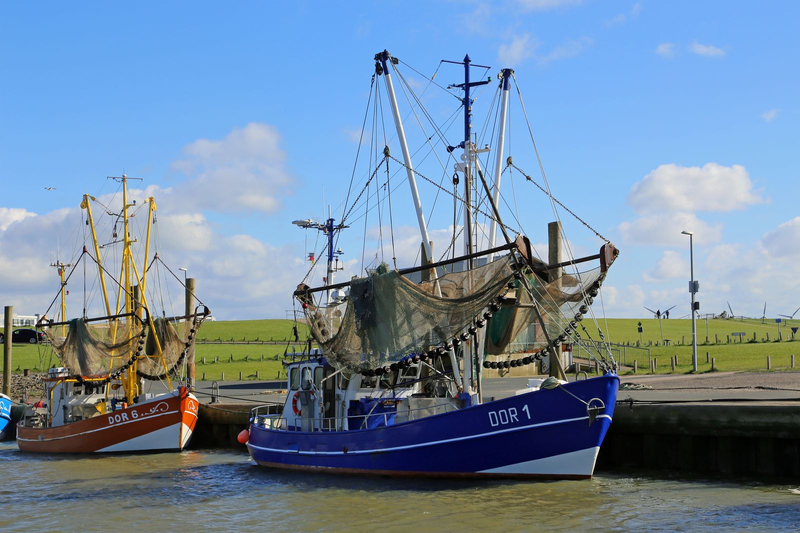 Two fishing boats with raised booms and nets moored at a quay wall, on the left an orange and white boat marked DOR 6, on the right a blue boat marked DOR 1, behind them a green dike under a blue sky with clouds