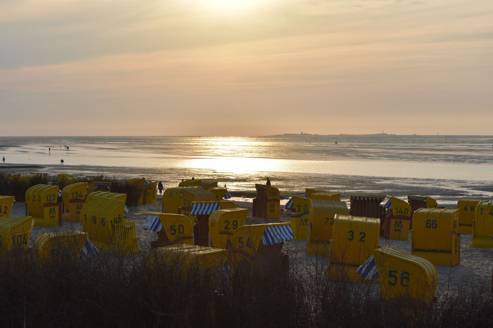 Weite Wattfläche bei Ebbe vor Cuxhaven-Duhnen bei Sonnenuntergang, im Vordergrund gelbe nummerierte Strandkörbe, am Horizont eine schmale Landsilhouette