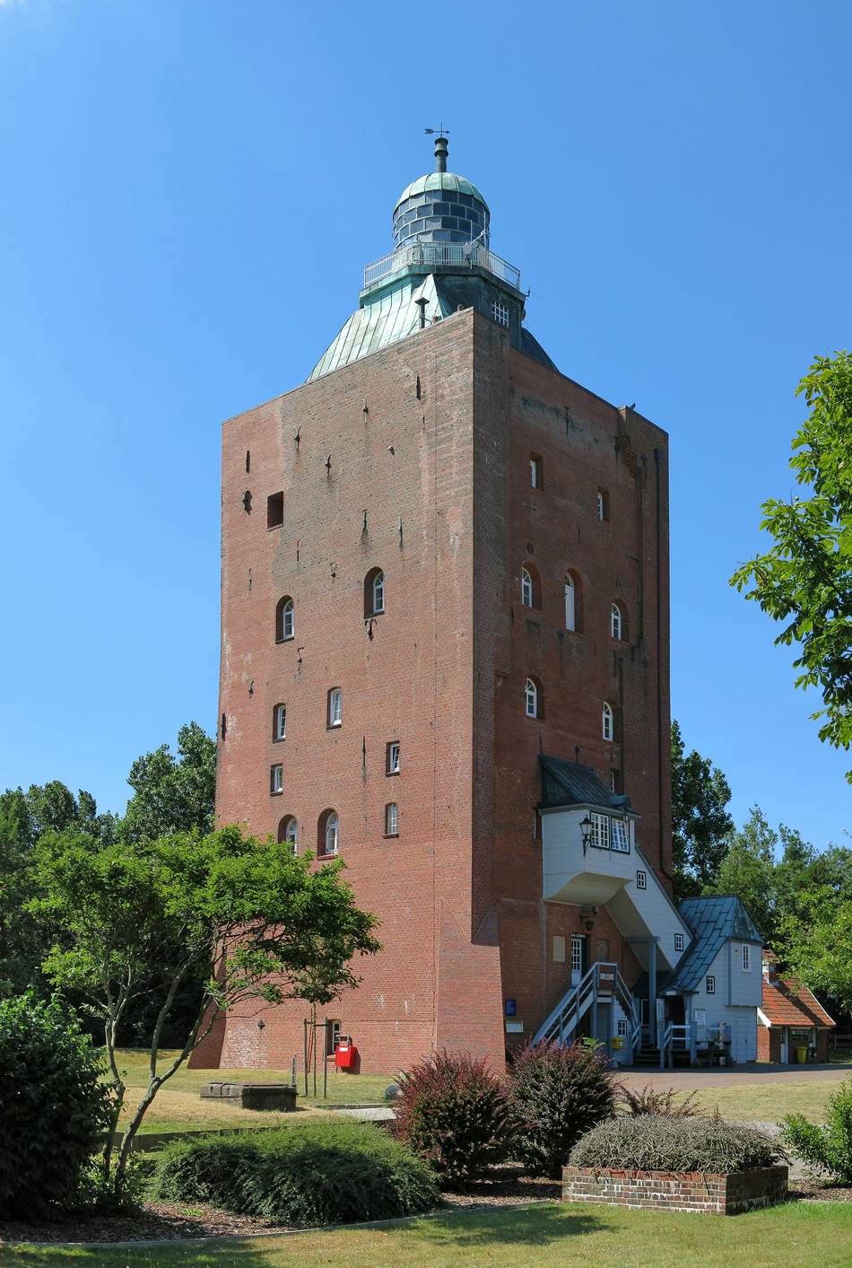 Backsteinturm des Leuchtturms Neuwerk mit grüner Laterne und weißem Anbau, umgeben von Bäumen und Rasen bei blauem Himmel
