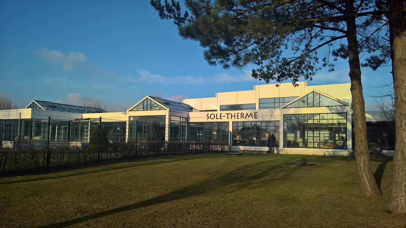 Low white hall building with large glass fronts and glass gabled roofs, the lettering SOLE-THERME above the glass front, a lawn with long shadows in front, a pine tree on the right, a fence with a hedge on the left, blue sky