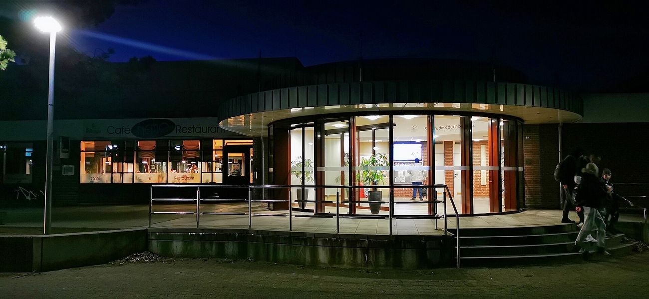 Round glazed entrance building in the dark, brightly lit inside with potted plants, a terrace with stainless steel railings and steps in front, a street lamp on the left and the lettering Café NEMO Restaurant above lit windows, a few people seen from behind on the right