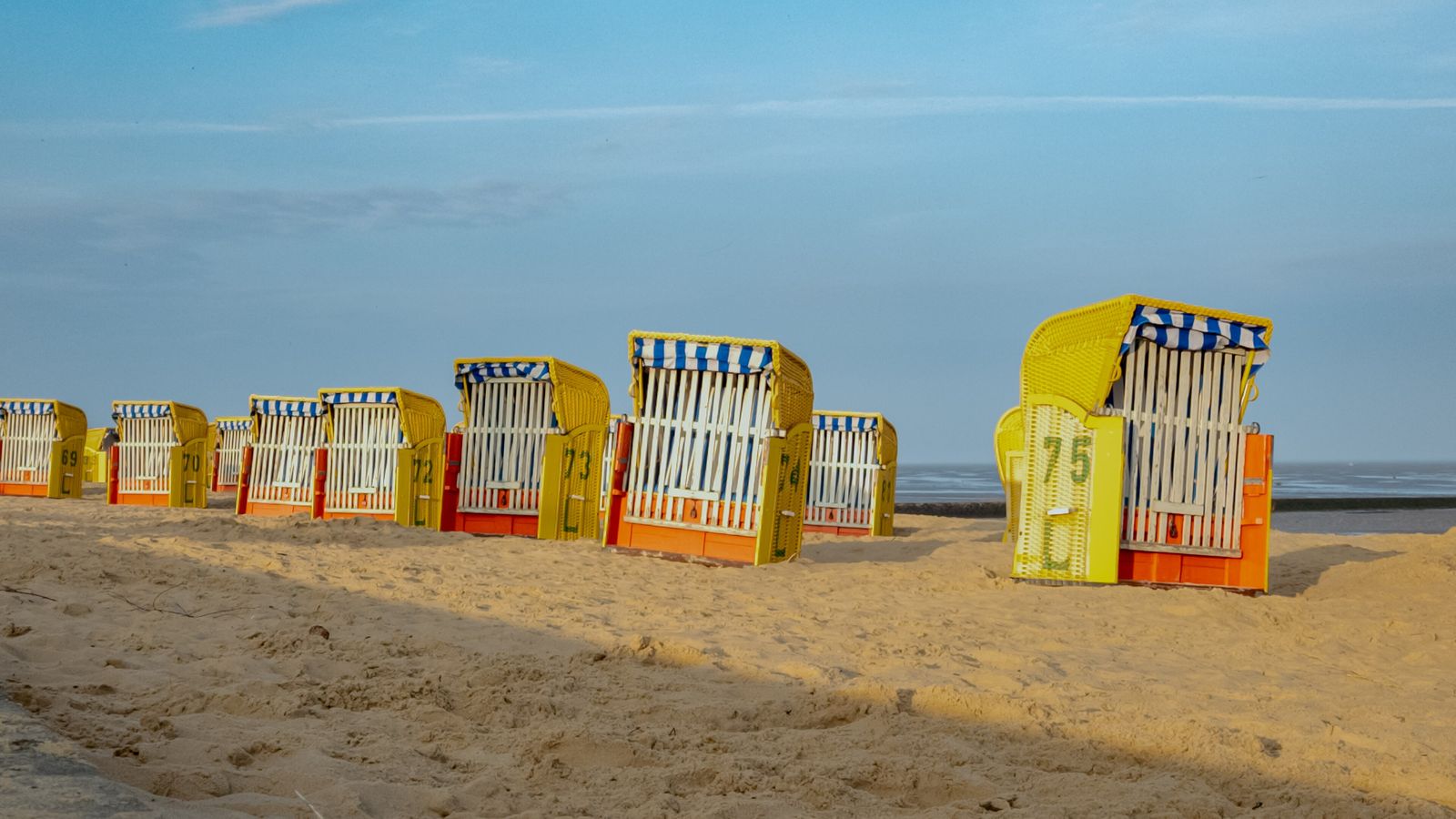 Row of yellow and white beach chairs with blue-and-white striped hoods on light sand, behind them a narrow strip of water under a blue sky