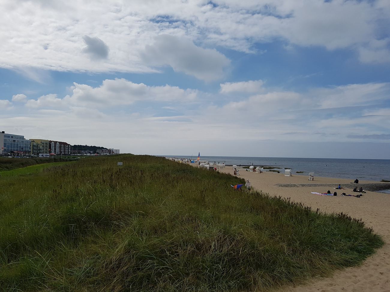 Grass-covered dune in the foreground, on the right a sandy beach with white beach chairs and a few people by the water, on the left in the distance multi-storey buildings, above them clouds and blue sky