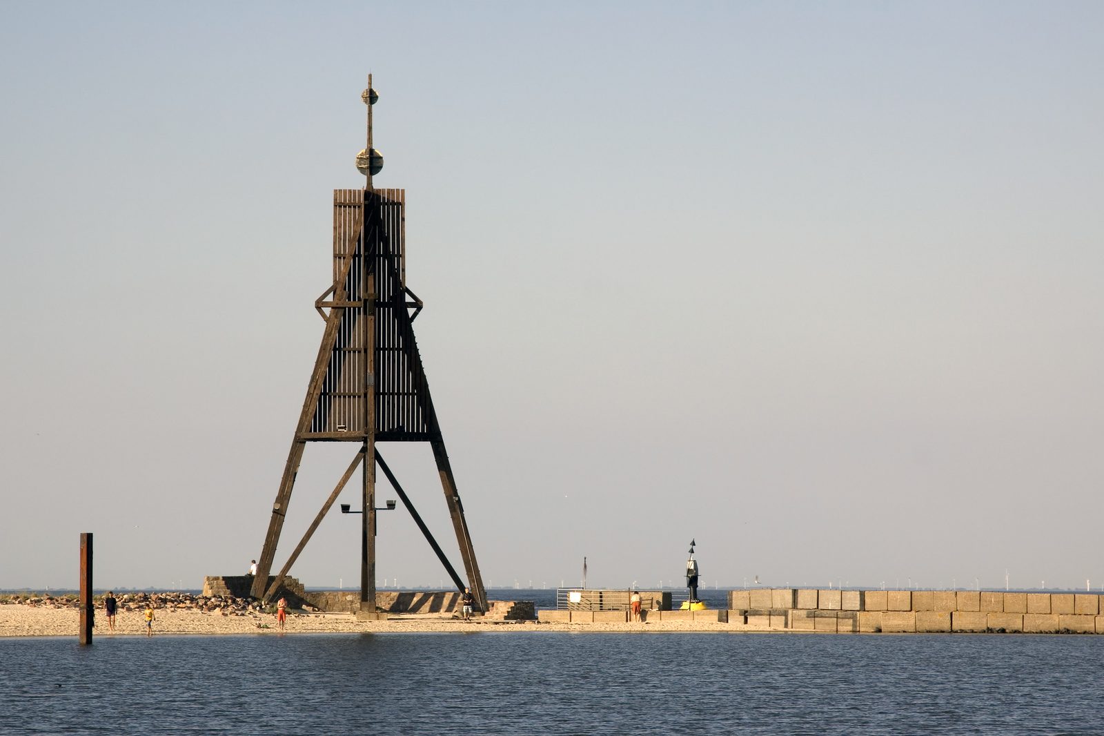 Hölzernes Seezeichen Kugelbake mit zwei Kugeln und Spitze auf einem Steinsockel am Strand, davor Wasser, am Horizont eine Mole und Windräder