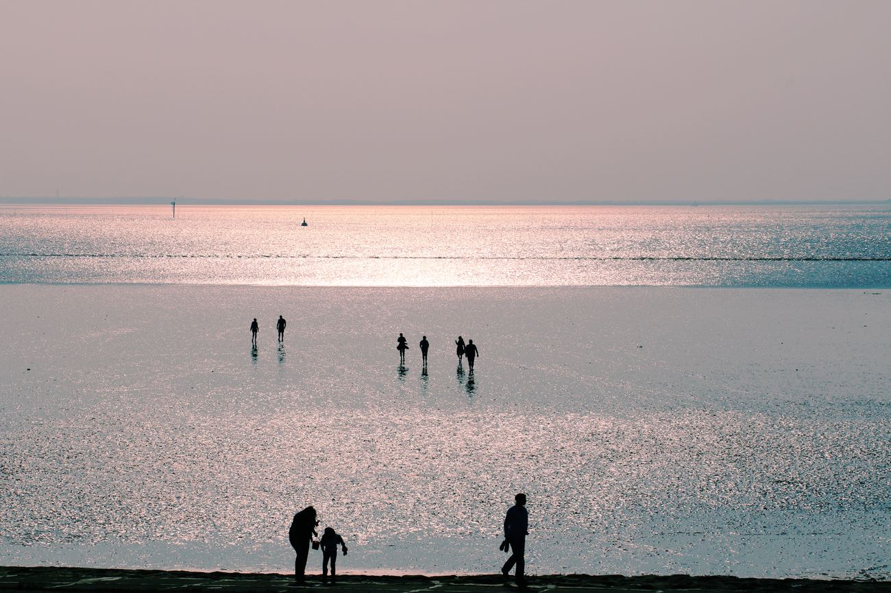 Silhouettes of several people walking across shimmering, water-covered mudflats in evening light (illustrative photo, not taken in Otterndorf)